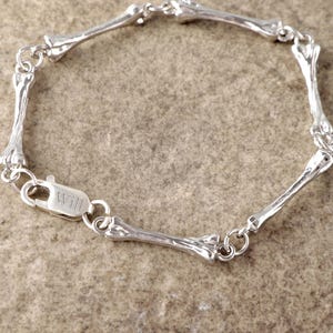May include: A silver-toned chain bracelet composed of six individual bone-shaped links connected by small circular jump rings. The bracelet features a lobster claw clasp with the word "Will" engraved on the surface of the metal closure mechanism.