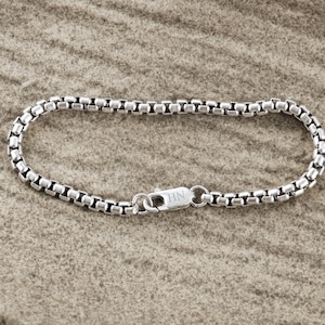Può includere: Un braccialetto a catena a scatola color argento con chiusura a moschettone. Il braccialetto è composto da piccole maglie rettangolari. La chiusura è incisa con le iniziali "HN". Il braccialetto è esposto su una superficie strutturata e neutra.