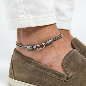 May include: A silver-colored anklet with an intricate braided design and a hook clasp. The anklet is worn on the ankle, showcasing the jewelry's details. The anklet is a stylish accessory.