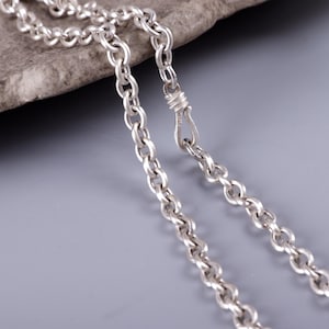 Op de afbeelding: Een zilveren ketting bestaande uit ronde, in elkaar grijpende schakels met een gepolijste metalen afwerking. De ketting is voorzien van een haakvormig sluitingsmechanisme dat de twee uiteinden verbindt tot een doorlopende lus van schakels.