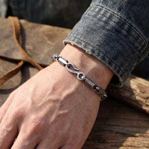 May include: Silver-toned bracelet with a hook clasp. The bracelet features rectangular links with engraved patterns. The hook clasp is a simple, curved design. The bracelet is worn on a wrist, with a denim jacket sleeve in the background.