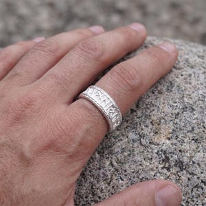 Può includere: Un anello a fascia color argento caratterizzato da una superficie testurizzata con caratteri runici incisi lungo l'intera circonferenza. Il metallo presenta una finitura opaca e una larghezza costante, con incisioni dettagliate.