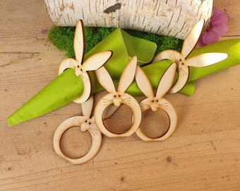 Puede incluir: Cuatro anillos de servilleta de madera en forma de conejo con una servilleta verde. Los anillos de conejo son de color marrón claro y tienen una cara blanca con ojos negros y bigotes.