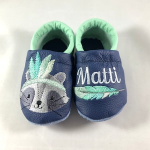 May include: A pair of blue leather baby moccasins with a raccoon wearing a headdress and a feather embroidered on one shoe and the name "Matti" embroidered on the other shoe.