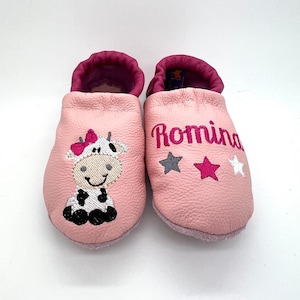 May include: Pink leather baby shoes with a cow embroidery and the name Romina embroidered on one shoe. The shoes have three silver stars and one pink star.