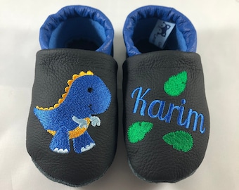 May include: Black leather baby shoes with blue trim. One shoe has a blue dinosaur with yellow accents embroidered on it. The other shoe has the name "Karim" embroidered on it in green.