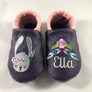 May include: Purple leather baby shoes with pink lining. One shoe has an embroidered bunny with a flower and feathers. The other shoe has the name "Ella" embroidered with flowers and feathers.
