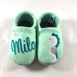 May include: A pair of light green baby moccasins with a seahorse and the name "Milo" embroidered on them.
