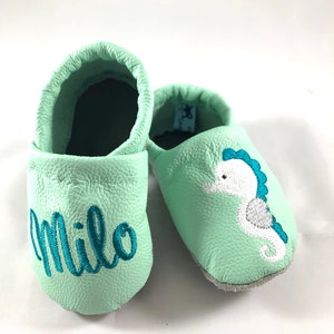 May include: A pair of light green baby moccasins with a seahorse design and the name "Milo" embroidered on one shoe.