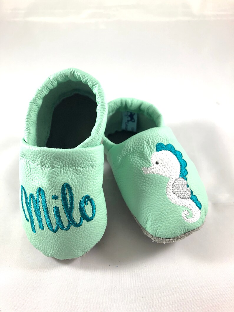 May include: A pair of light green baby moccasins with a seahorse design and the name "Milo" embroidered on one shoe.