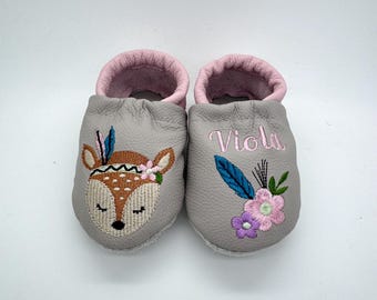 May include: A pair of gray baby moccasins with pink lining. One shoe features an embroidered deer face with a floral crown, the other has the name "Viola" with floral embroidery. The shoes are designed for infants.