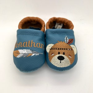 May include: A pair of blue leather baby moccasins with brown leather trim. One moccasin has the name "Jonathan" embroidered on it, and the other has a brown bear with a feather headdress embroidered on it.