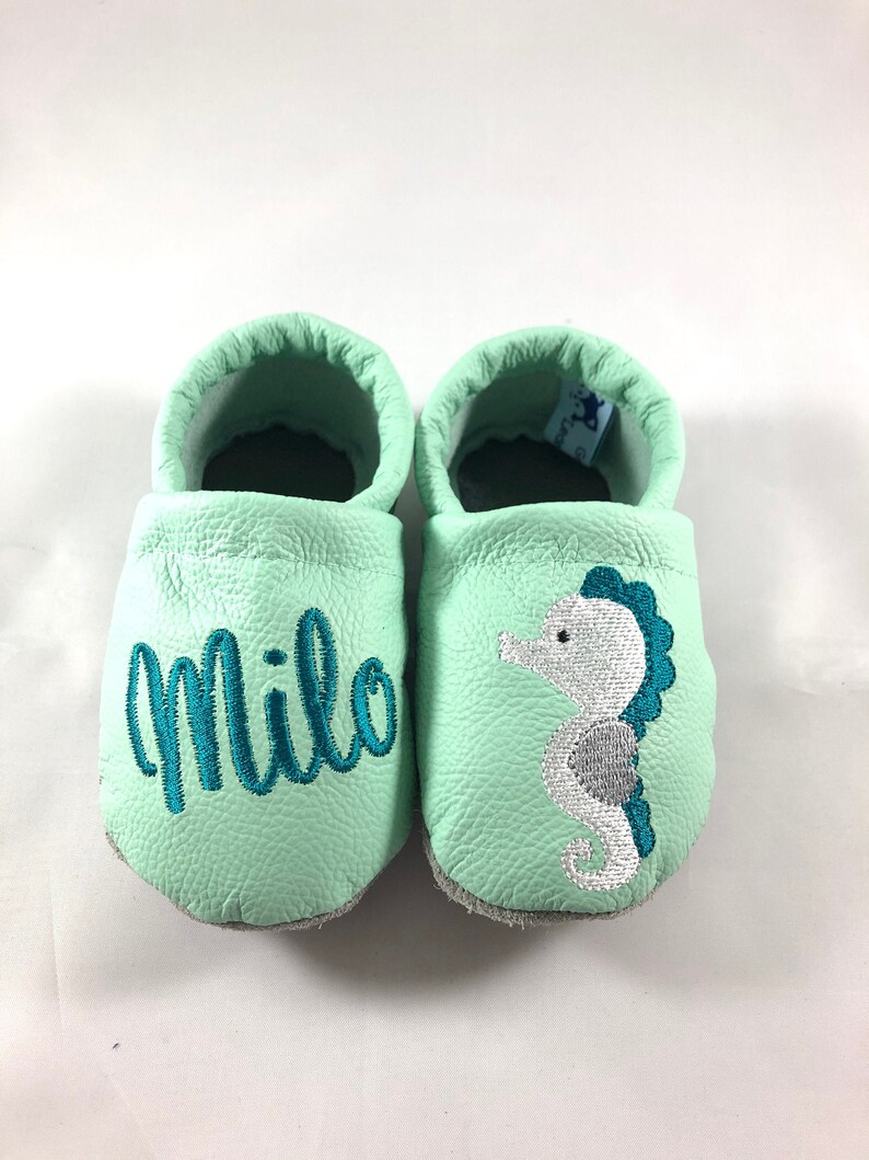 May include: A pair of light green baby moccasins with a seahorse and the name "Milo" embroidered on them.