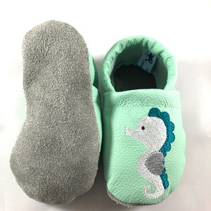 May include: A pair of light green leather baby moccasins with a grey suede sole. The moccasins have a white embroidered seahorse with teal accents.