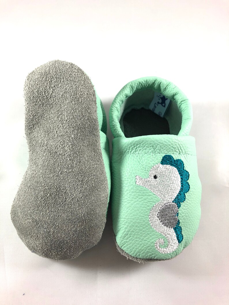 May include: A pair of light green leather baby moccasins with a grey suede sole. The moccasins have a white embroidered seahorse with teal accents.