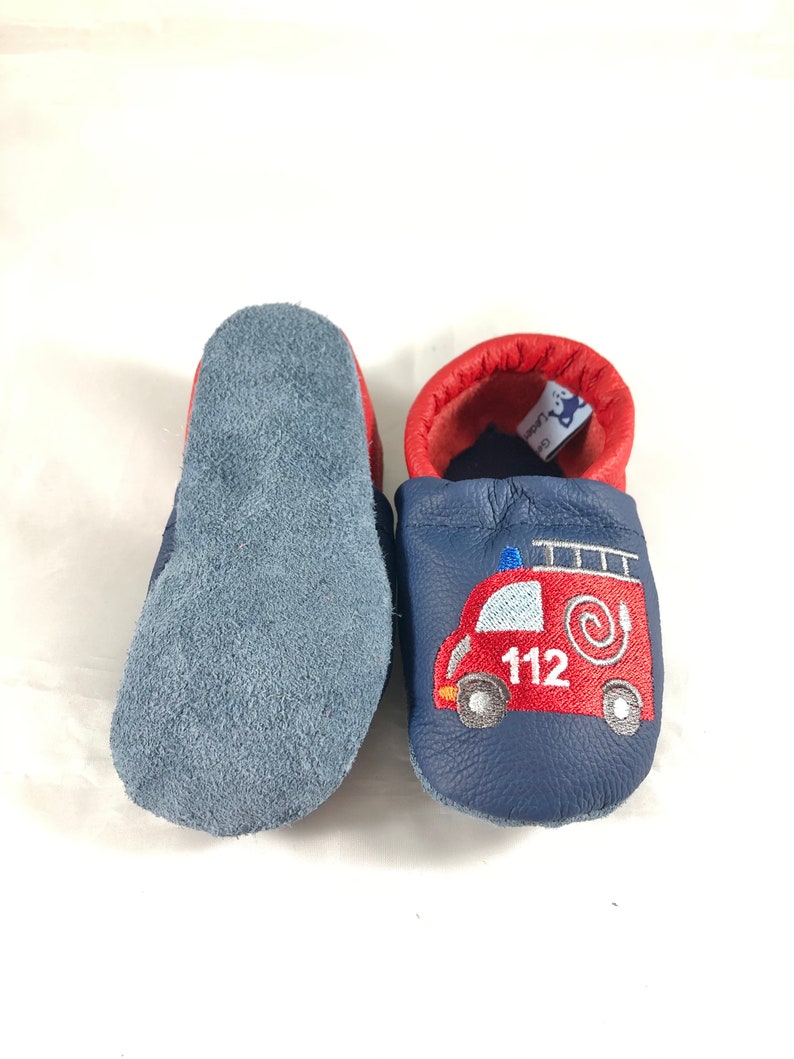 May include: A pair of blue leather baby shoes with a red fire truck embroidered on one shoe. The shoes have a soft suede sole.