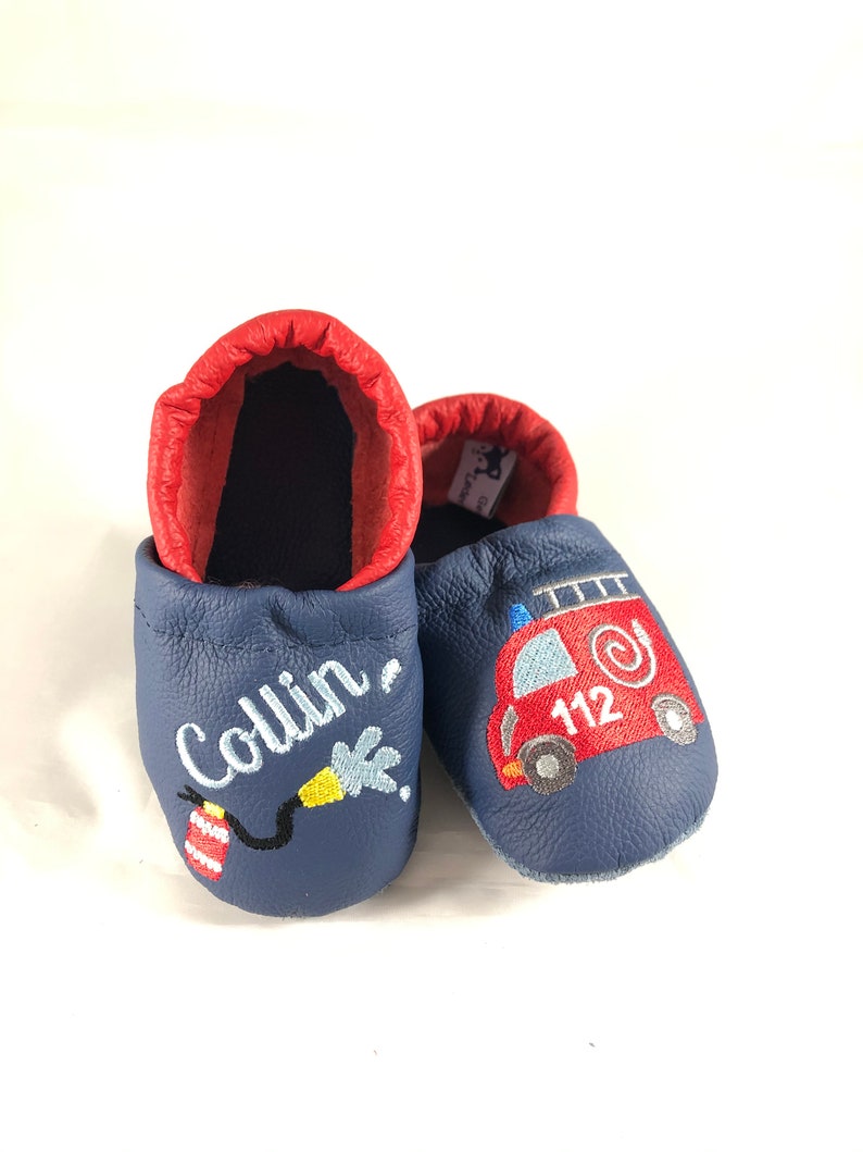 May include: Blue leather baby shoes with red trim. One shoe has the name "Collin" embroidered on it, and the other shoe has a red fire truck with the number "112" embroidered on it.