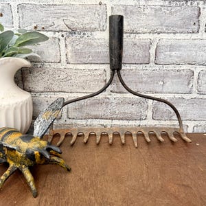 May include: A rustic garden rake with a dark wooden handle and metal tines. The rake is set against a brick wall and a wooden surface. A decorative bee figurine and a white vase are also in the image.
