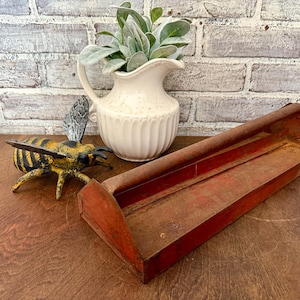 May include: A decorative bee figurine with yellow and black stripes, next to a white pitcher with faux greenery and a red metal tray. The pitcher has a textured surface and a curved handle. The tray has a rectangular shape.