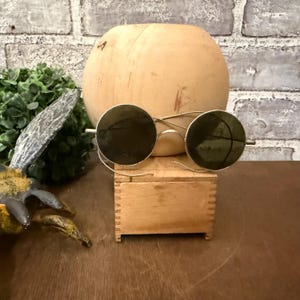 May include: Vintage round sunglasses with gold frames and dark lenses. The sunglasses rest on a small wooden box, with a wooden hat form in the background. The background includes a brick wall and greenery.