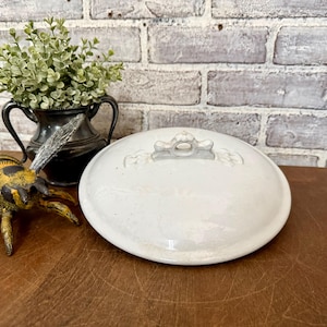 May include: A white ceramic serving dish with a lid. The lid has a decorative handle and embossed floral details. The dish is on a wooden surface, with a decorative metal vase and a bee figurine in the background.