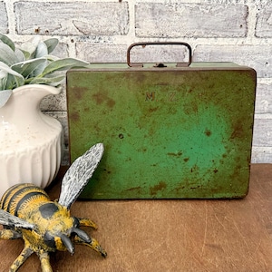 May include: A rectangular green metal lunch box with a dark metal handle and latch. The surface shows signs of wear and rust, with the letters M. Z. engraved on the front. The box is positioned next to a white vase and a bee figurine.