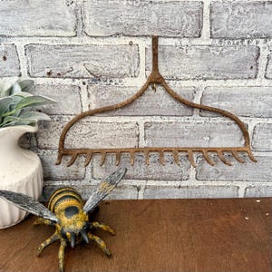 May include: A vintage metal rake with a curved handle and tines, displayed against a distressed brick wall. A decorative bee figurine with yellow and black stripes is in front of the rake. A white vase with green leaves is on the left.