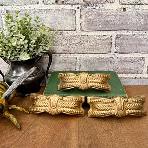 May include: Three gold-colored, decorative bow-shaped drawer pulls with an ornate design. The pulls are arranged on a wooden surface with a green book and a brick background. A decorative metal pitcher and a bee figurine are also visible.