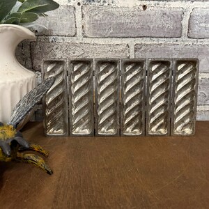May include: A vintage, silver-colored metal mold with six rectangular compartments. Each compartment features a twisted, rope-like design. The mold is likely used for baking or creating decorative items. The background includes a weathered brick wall and a wooden surface.