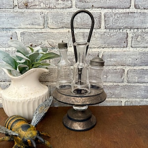 May include: A vintage-style condiment set with three clear glass bottles and a metal stand. The bottles have metal lids and a central glass bottle with a stopper. The stand is round with a handle.