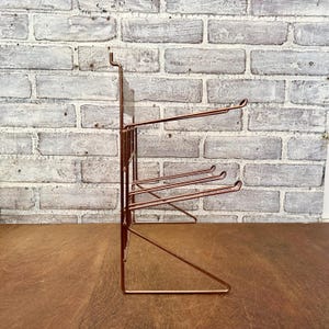 May include: A copper-colored metal wall-mounted organizer with multiple horizontal bars for hanging items. The organizer has a rectangular frame and is designed to be mounted on a wall. The background is a brick wall.