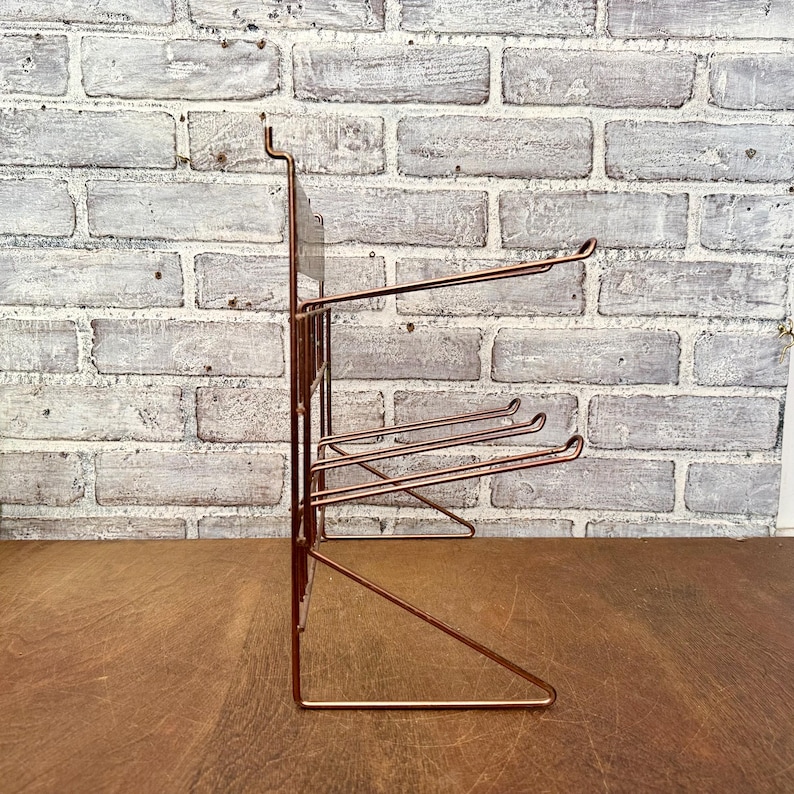 May include: A copper-colored metal wall-mounted organizer with multiple horizontal bars for hanging items. The organizer has a rectangular frame and is designed to be mounted on a wall. The background is a brick wall.