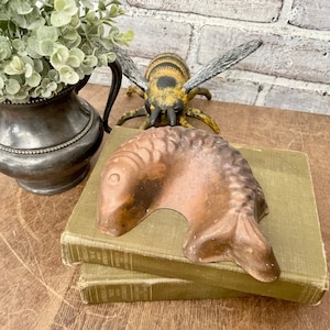 May include: A brown ceramic fish-shaped dish sits on top of two green books. A black and yellow toy bee is in the background.