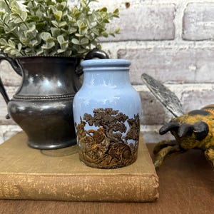 May include: A pale blue ceramic vase featuring a brown relief design of figures and trees. The vase has a cylindrical shape with a slightly flared top and rests on a vintage book. A silver jug and decorative bee are in the background.