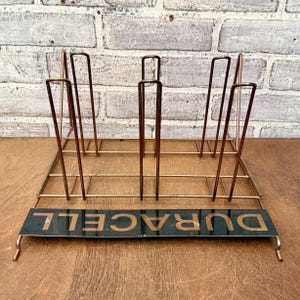 May include: A vintage copper-toned metal rack with vertical dividers and a rectangular base. The base features the word "DURACELL" in gold lettering on a black background. The rack is designed to hold items upright.