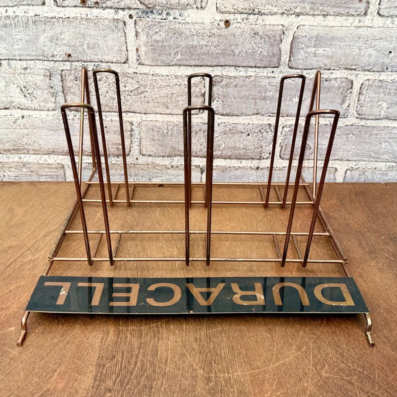 May include: A vintage copper-toned metal rack with vertical dividers and a rectangular base. The base features the word "DURACELL" in gold lettering on a black background. The rack is designed to hold items upright.