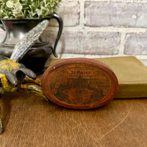 May include: An antique, oval-shaped, wooden box with a faded red finish. The box features an image of a building and the text "St Peter, North Tichill, R.A. Foley". The box is resting on a wooden surface.