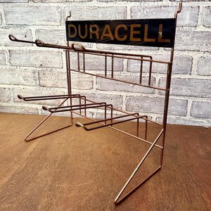 May include: Vintage copper-toned wire display rack with the word "DURACELL" in gold lettering on a black sign. The rack has multiple shelves and angled supports, ideal for displaying small items. The rack is set against a brick wall.