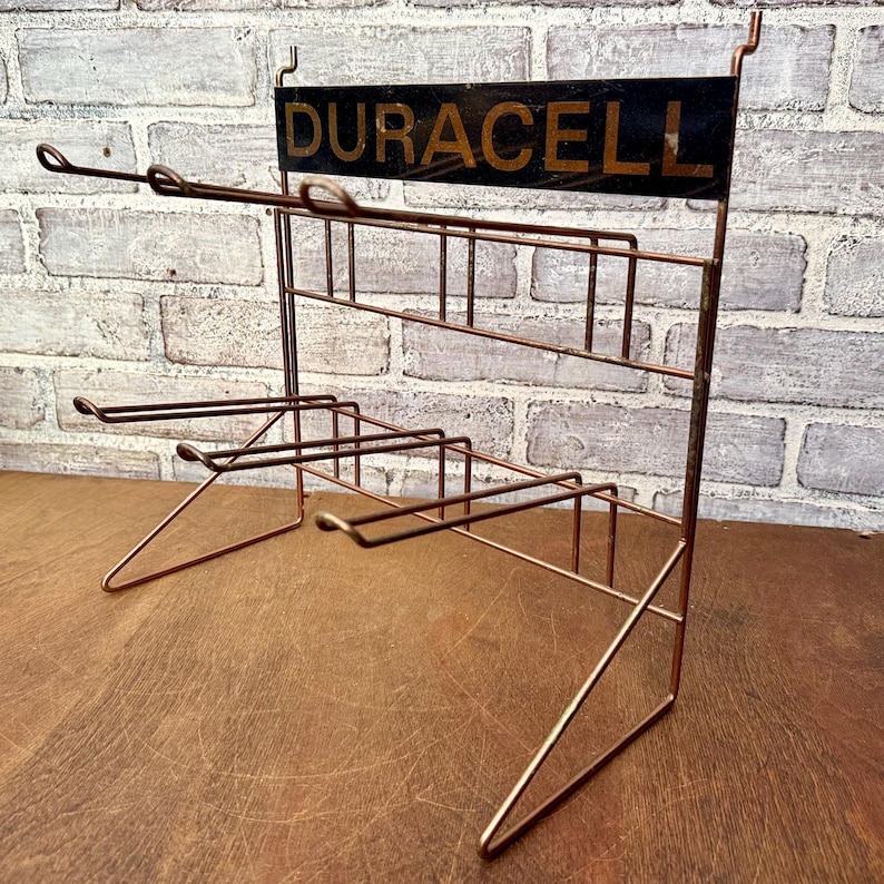 May include: Vintage copper-toned wire display rack with the word "DURACELL" in gold lettering on a black sign. The rack has multiple shelves and angled supports, ideal for displaying small items. The rack is set against a brick wall.