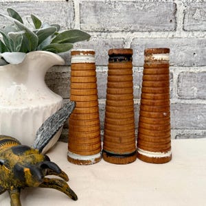 May include: Three cylindrical wooden spools with horizontal ridges, featuring natural brown wood tones and distressed white or black painted accents at the top and bottom edges, arranged vertically in a row on a flat surface.