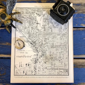 May include: Vintage map of Seattle, Washington, with a compass, a toy bee, and a vintage camera on a blue wooden surface. The map shows the city's streets and landmarks, including Elliott Bay.