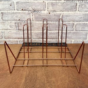 May include: A copper-colored metal record holder with a rectangular base and vertical dividers. The holder has a geometric design with angled supports on each end. The word "DURACELL" is visible on a black strip at the base.
