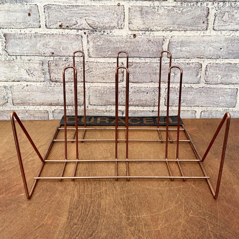May include: A copper-colored metal record holder with a rectangular base and vertical dividers. The holder has a geometric design with angled supports on each end. The word "DURACELL" is visible on a black strip at the base.
