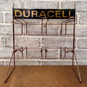 May include: A vintage copper-colored metal battery display rack with the word "DURACELL" in gold lettering on a black background. The rack has multiple shelves and hooks for displaying batteries. The rack is set against a brick wall.