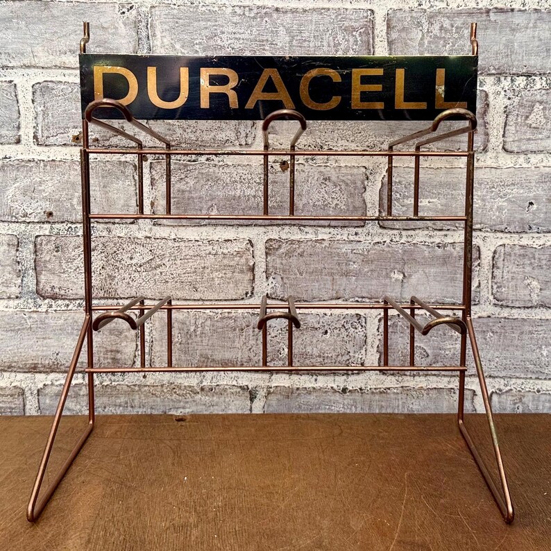 May include: A vintage copper-colored metal battery display rack with the word "DURACELL" in gold lettering on a black background. The rack has multiple shelves and hooks for displaying batteries. The rack is set against a brick wall.