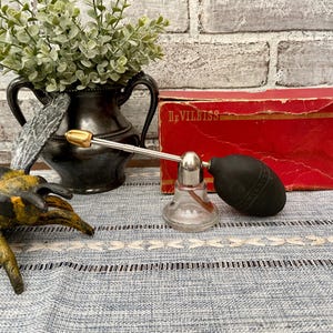 May include: An antique perfume atomizer with a clear glass bottle, silver-colored metal accents, and a black bulb. A red box with the text "DeVilbiss" is behind the atomizer. A decorative black vase with greenery and a metal bee figurine are also present.