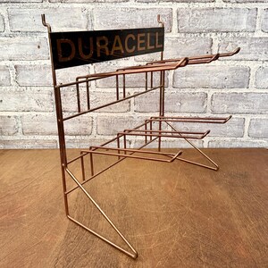 May include: A vintage copper-toned wire display rack with multiple shelves. The top features a black sign with the word "DURACELL" in gold lettering. The rack is designed to hold and display products, likely batteries.
