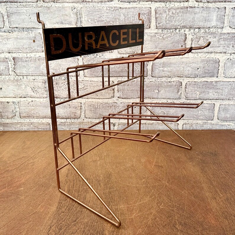 May include: A vintage copper-toned wire display rack with multiple shelves. The top features a black sign with the word "DURACELL" in gold lettering. The rack is designed to hold and display products, likely batteries.