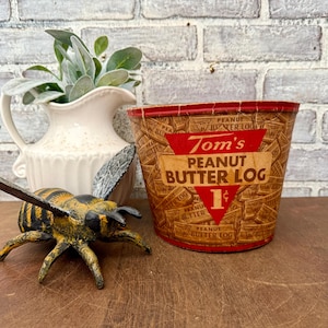 May include: Vintage Tom's Peanut Butter Log container with a red and brown design. The container has the text "Tom's PEANUT BUTTER LOG 1¢". A decorative bee and a white pitcher with greenery are also in the image.