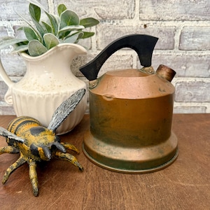 May include: A vintage copper kettle with a black handle and spout, next to a decorative bee figurine and a white pitcher with greenery. The kettle has a round base and a slightly aged appearance, suggesting it's an antique.